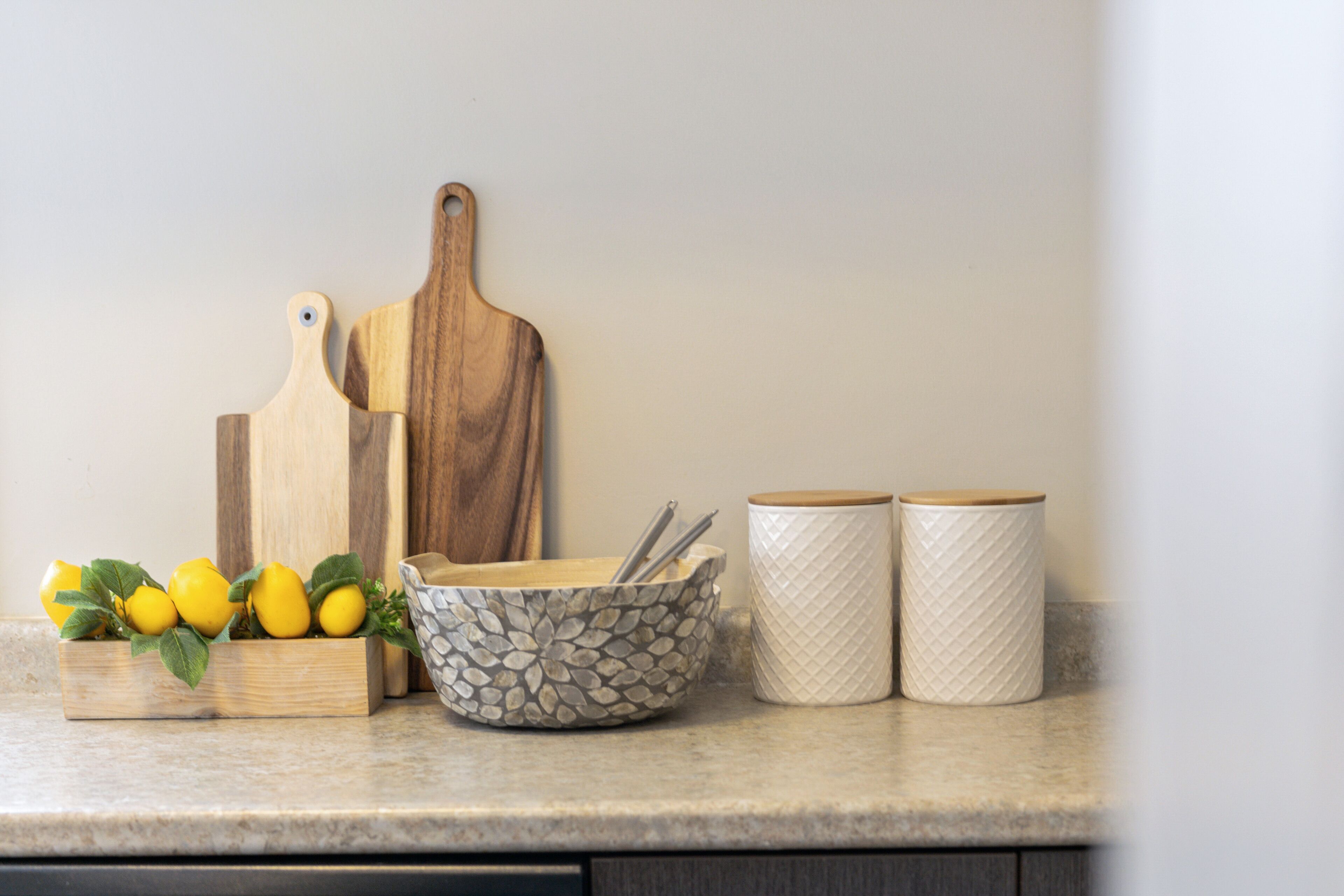 A kitchen counter with a wooden cutting board, a patterned bowl, two white containers, and a wooden box with lemons.