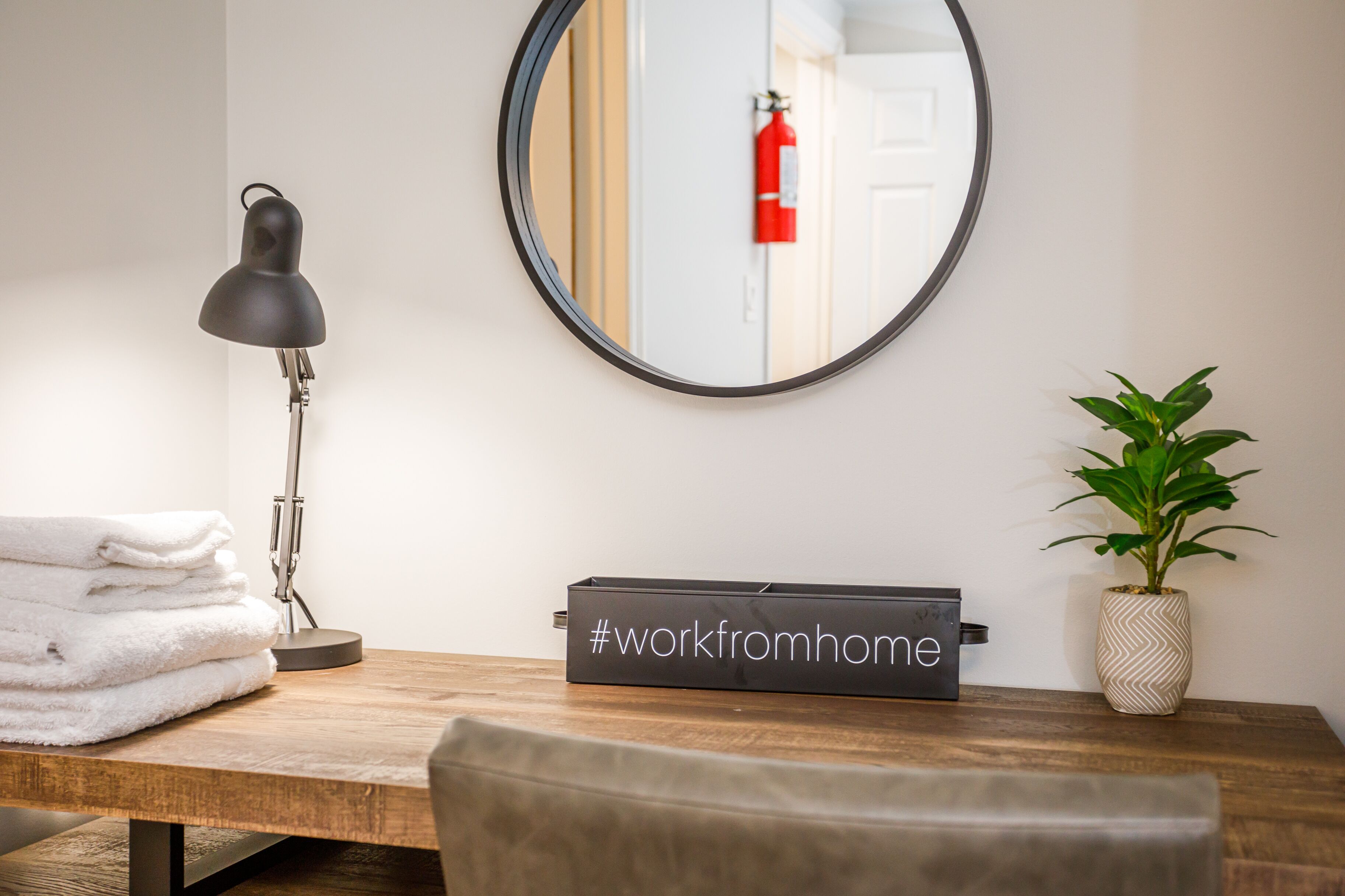A neatly arranged workspace with a desk, a lamp, a stack of towels, a potted plant, and a mirror reflecting a door and a fire extinguisher.