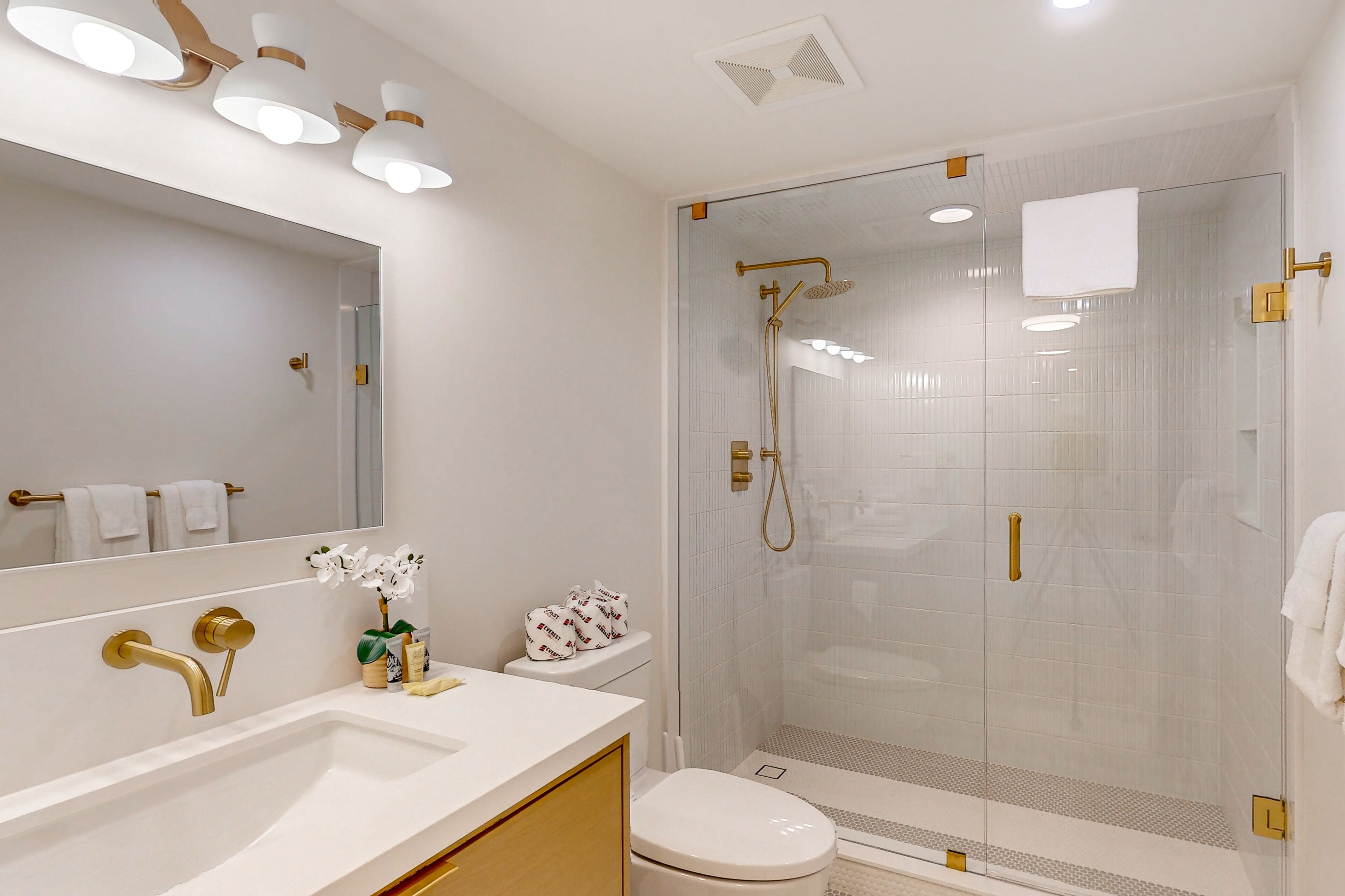 A modern bathroom with a large mirror, a white sink with gold fixtures, a toilet, and a glass-enclosed shower area.