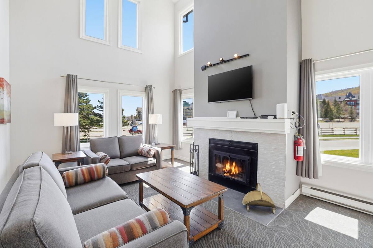 A modern living room with a fireplace, a flat-screen TV mounted above it, and a large window offering a view of the outdoors.