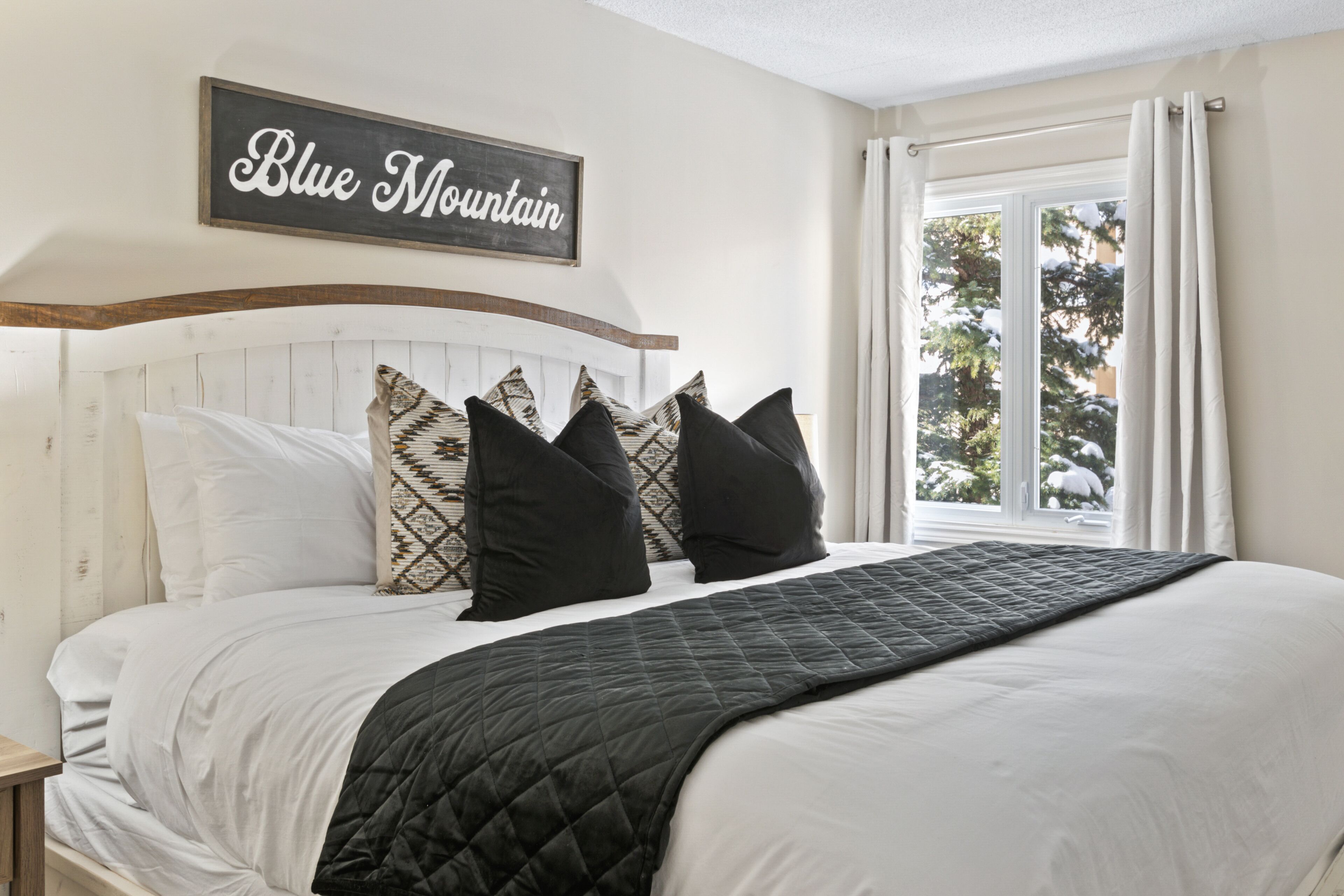 A bedroom with a bed, pillows, a quilted blanket, a headboard, a window with curtains, and a sign on the wall.