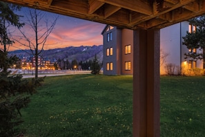 A building with multiple windows, a grassy area with dandelions, and a view of a distant town with lights.
