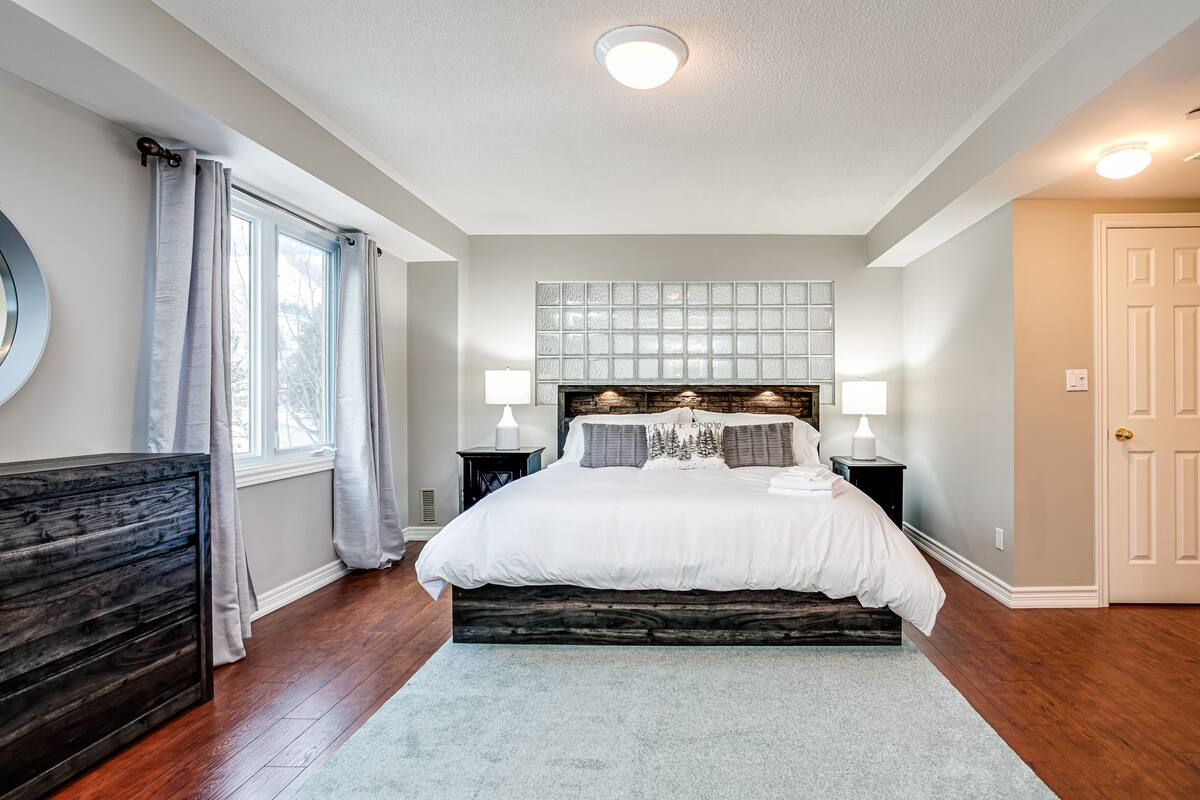A bedroom with a large bed, a wooden headboard, a nightstand with a lamp, a dresser, and a window with curtains.