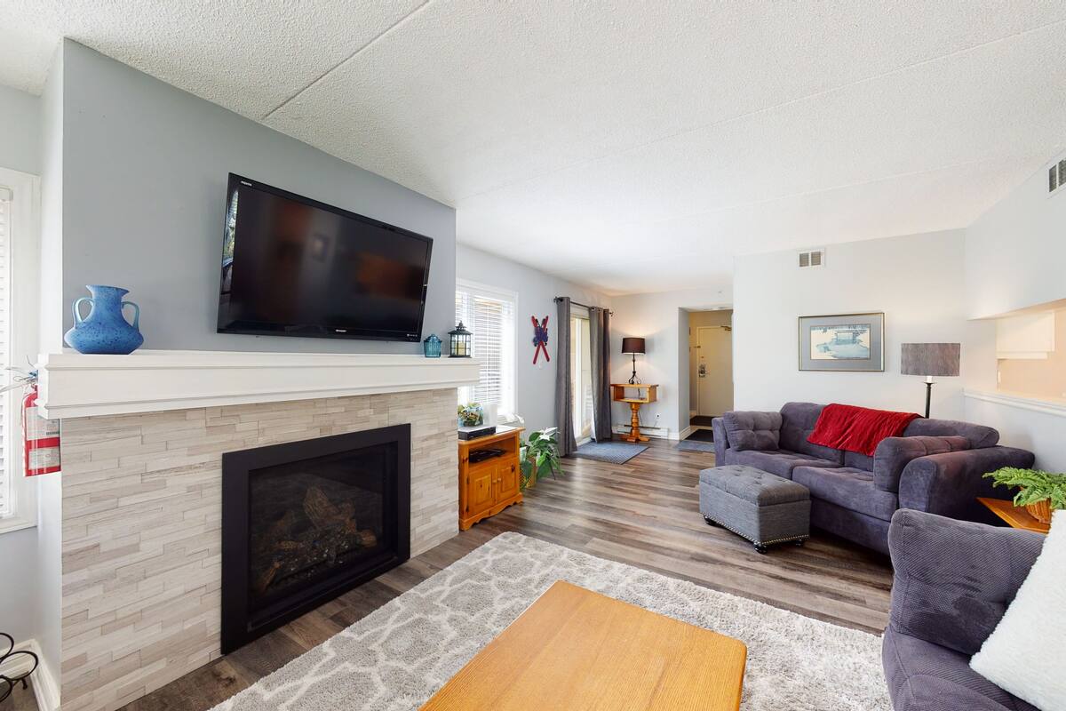 A living room with a fireplace, a flat-screen TV mounted above it, a grey sofa, and a wooden coffee table.