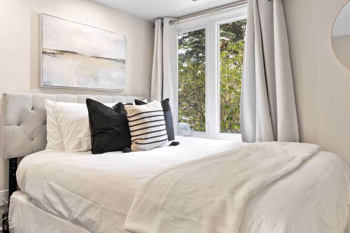 A bedroom with a bed, a headboard, a window with curtains, and a wall-mounted artwork.