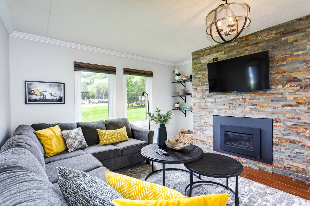A modern living room with a grey sofa, yellow cushions, a round coffee table, and a fireplace with a TV above it.