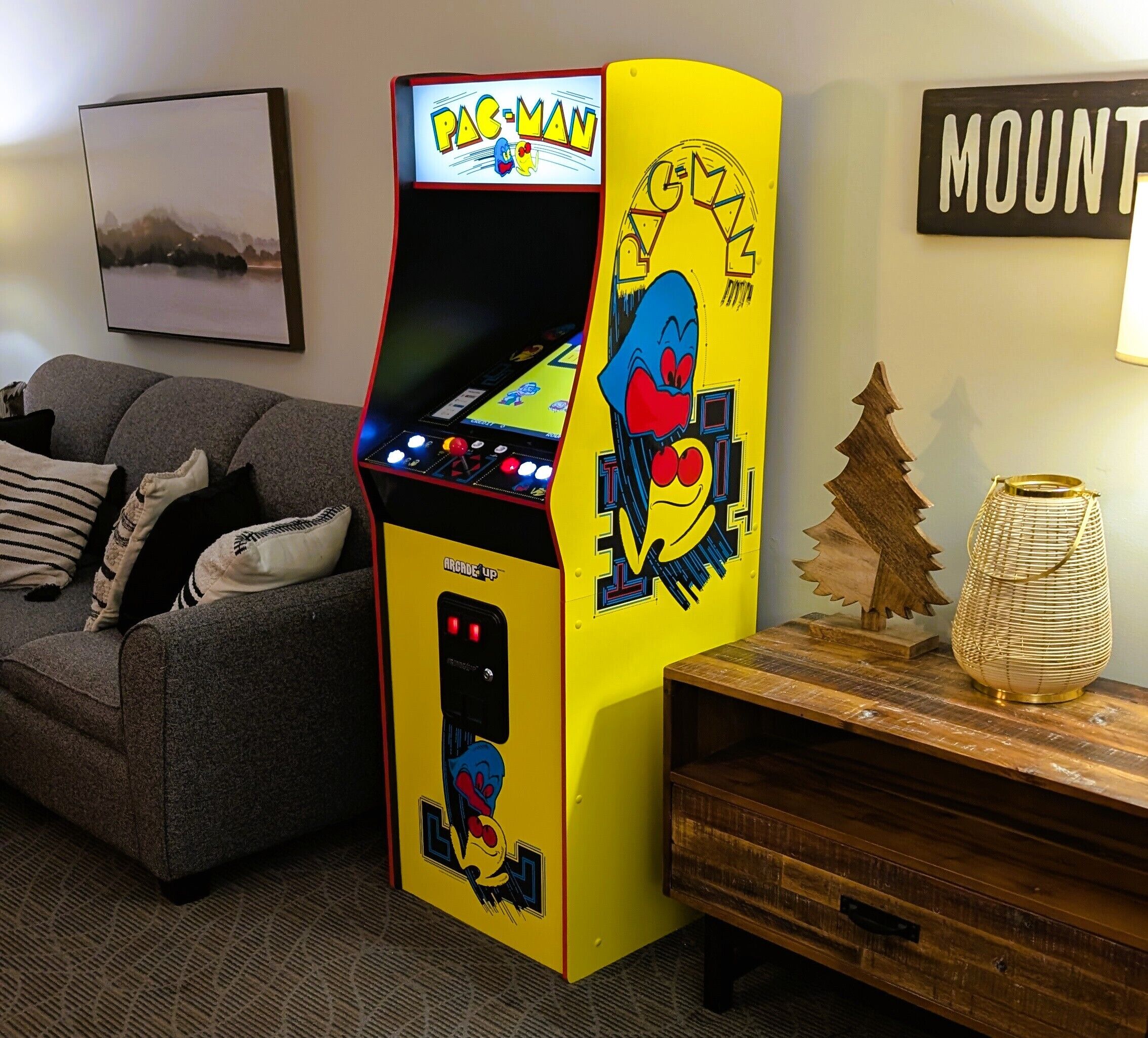 A Pac-Man arcade machine in a living room with a couch, a wooden side table, and a framed picture on the wall.