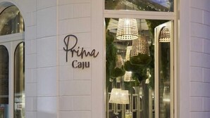 A restaurant entrance with a sign reading "Prima Caju," featuring a modern interior with hanging lamps and a visible dining area.
