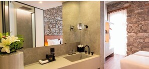 A modern bathroom with a stone wall, a large mirror, a sink with toiletries, and a bed with a stone wall backdrop. - Caju Le Petit Hotel (Funchal)
