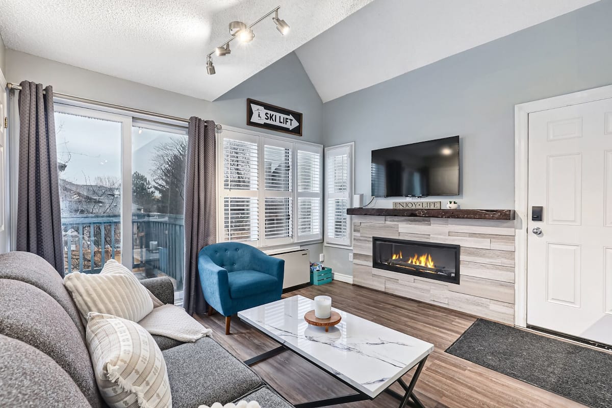 Una sala de estar con sofá, un sillón azul, una chimenea y un televisor montado en la pared.