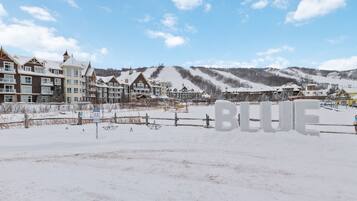 A snow-covered landscape with a row of multi-story buildings, likely a hotel or resort, with a clear sky and distant hills.