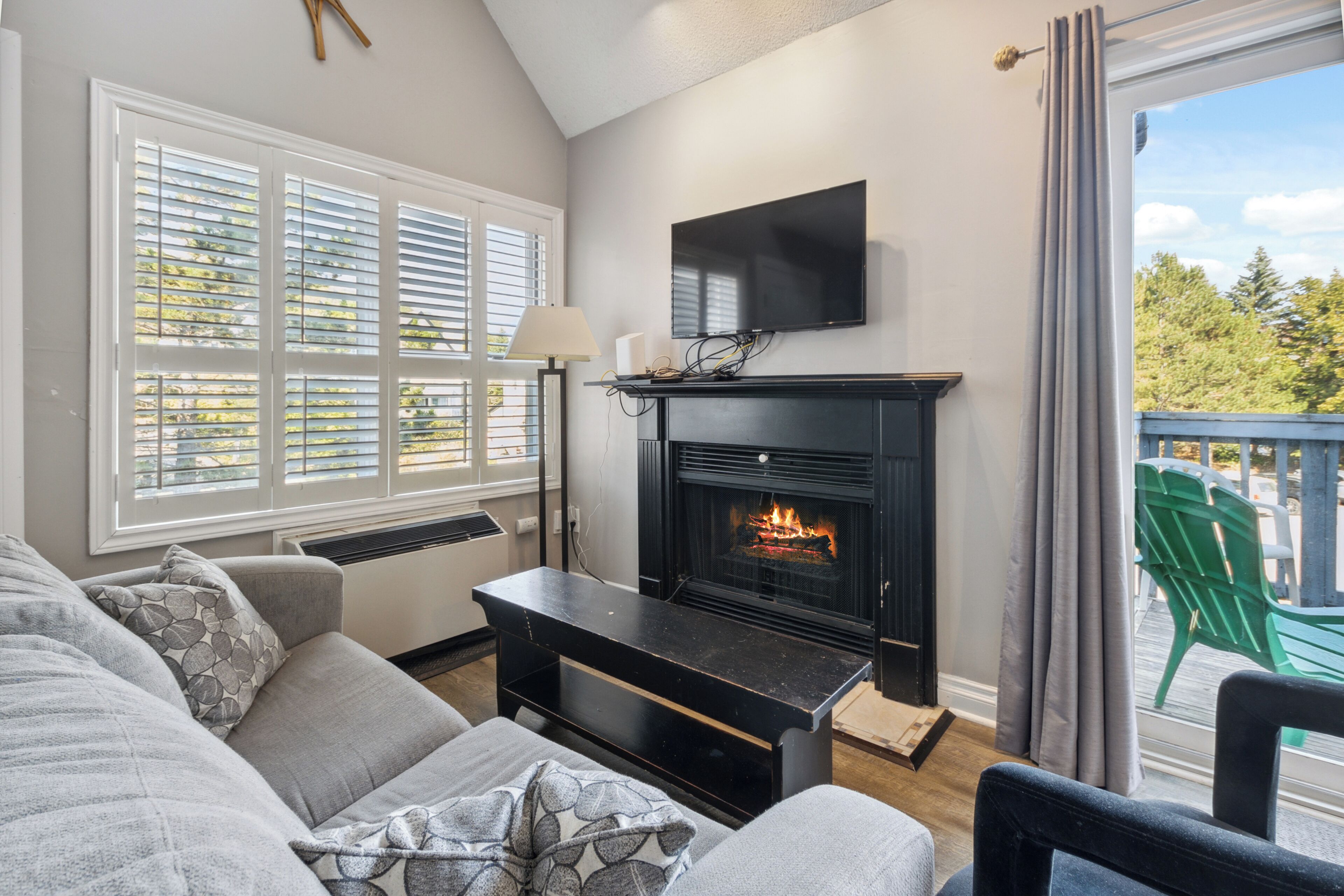 A living room with a fireplace, a television mounted above it, and a view of the outdoors through large windows.