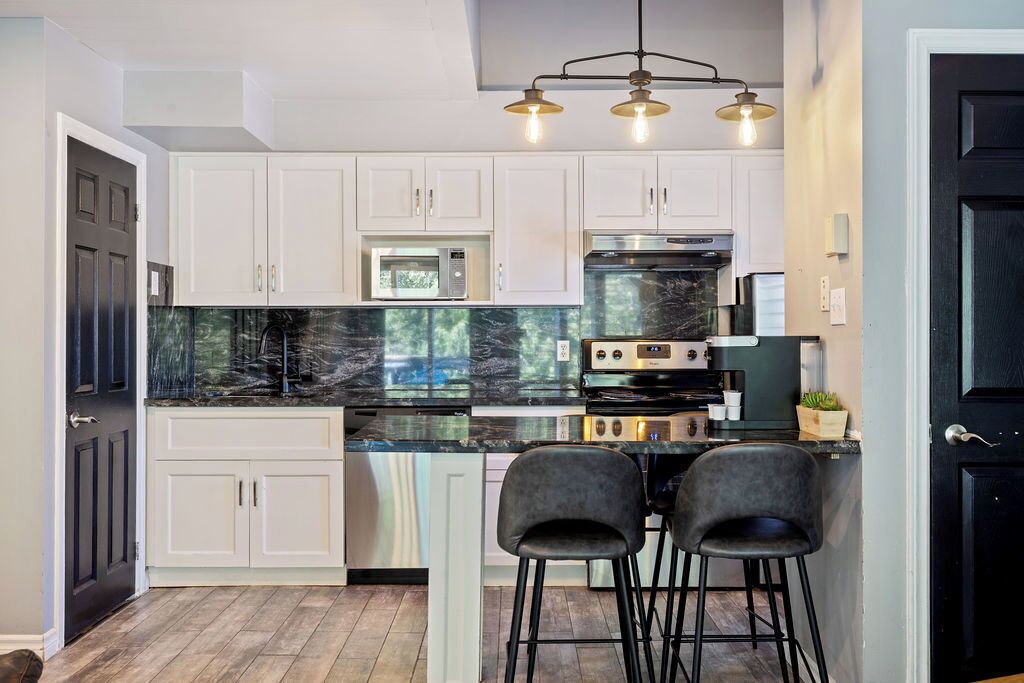 A modern kitchen with white cabinets, a black marble countertop, and a built-in oven. There are two black bar stools and a pendant light fixture above the counter.