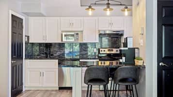 A modern kitchen with white cabinets, a black marble countertop, and a built-in oven. There are two black bar stools and a pendant light fixture above the counter.