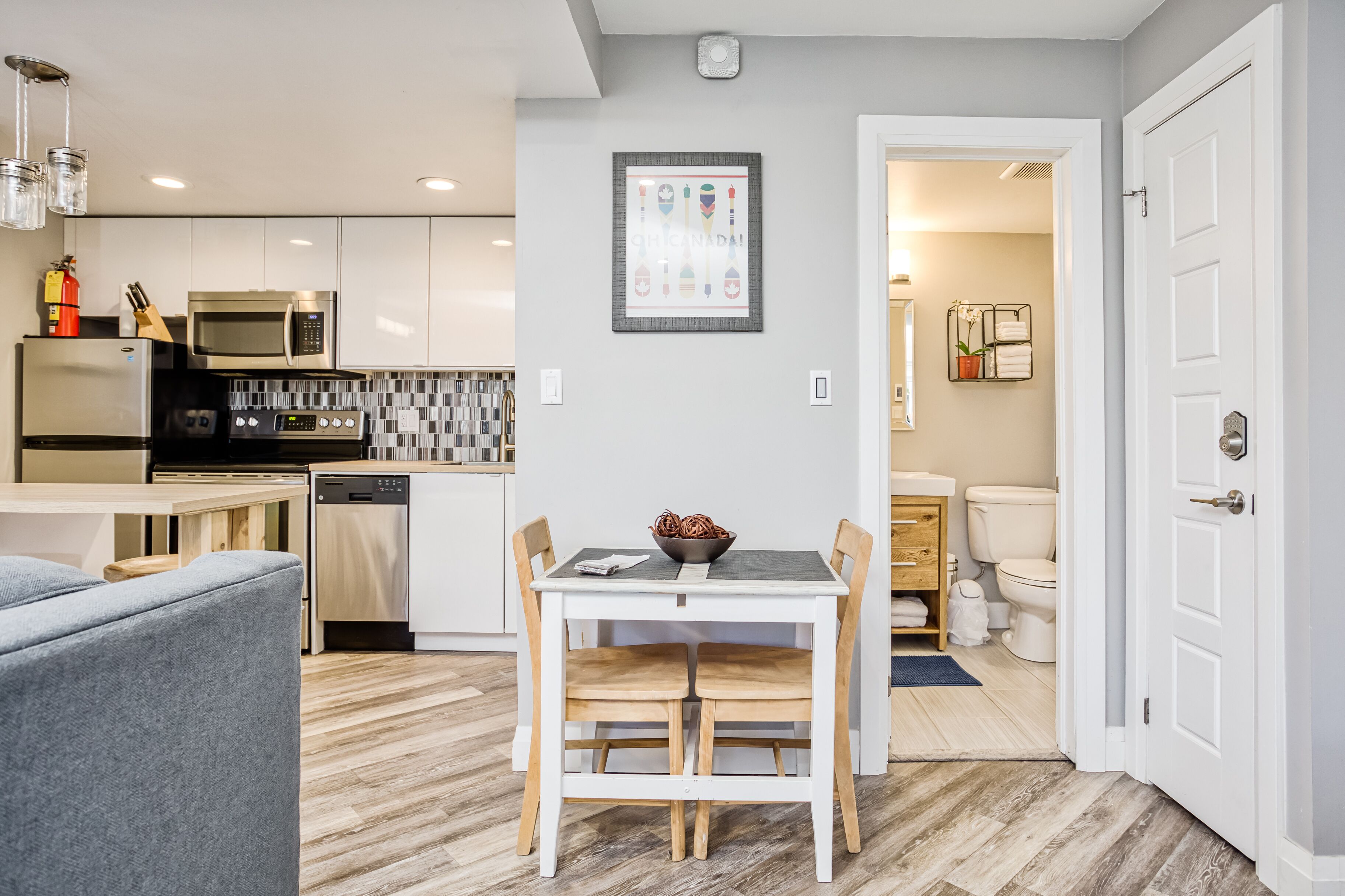 A modern kitchen with stainless steel appliances, a dining area with a white table and wooden chairs, and a visible bathroom with a toilet and sink.