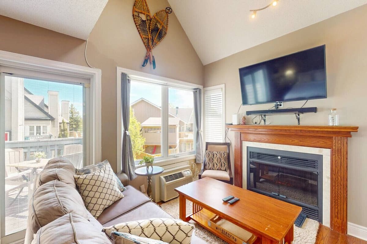 Una sala de estar con sofá, mesa de centro, chimenea, televisor y ventanas amplias.