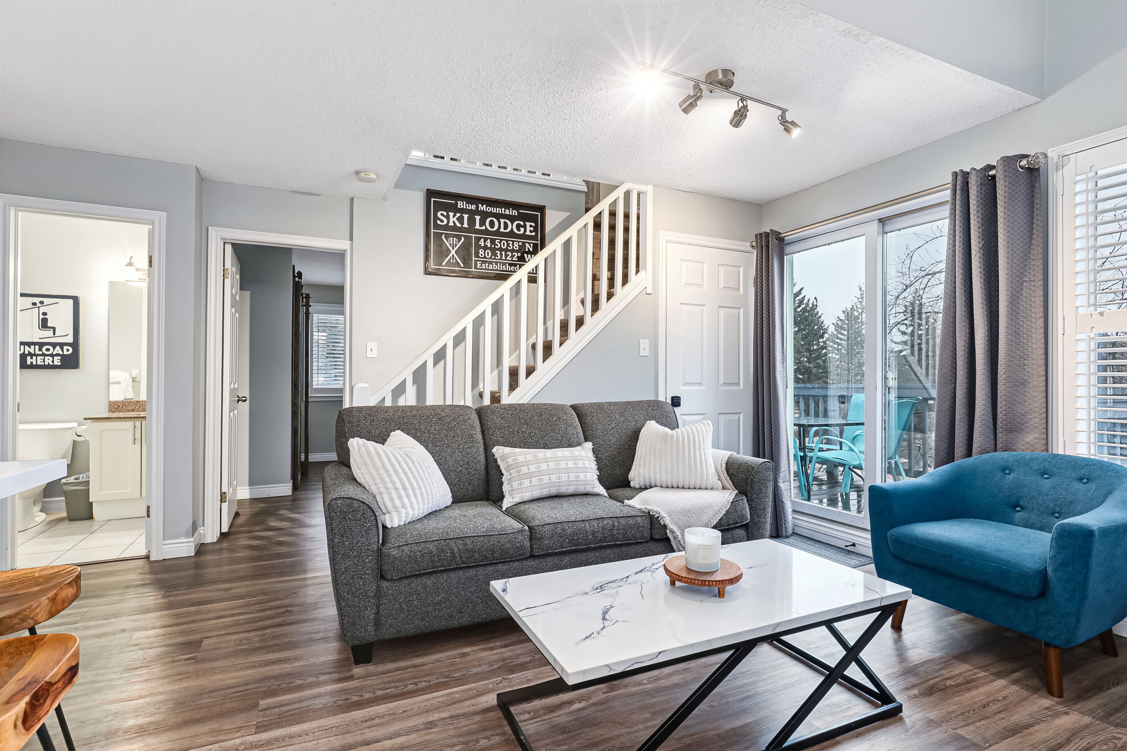 A modern living room with a grey sofa, a blue armchair, and a white coffee table. A staircase leads to a balcony with a view of trees.