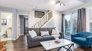 A modern living room with a grey sofa, a blue armchair, and a white coffee table. A staircase leads to a balcony with a view of trees.