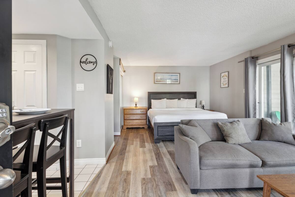 Un dormitorio moderno con cama, sofá gris, mesita de noche de madera y escritorio con sillas.