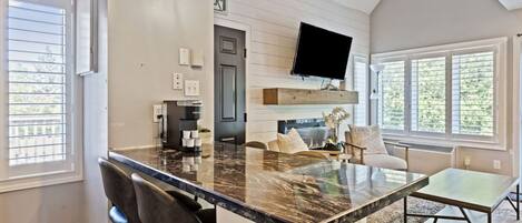 A modern kitchen with a marble island, bar stools, a flat-screen TV, and large windows.