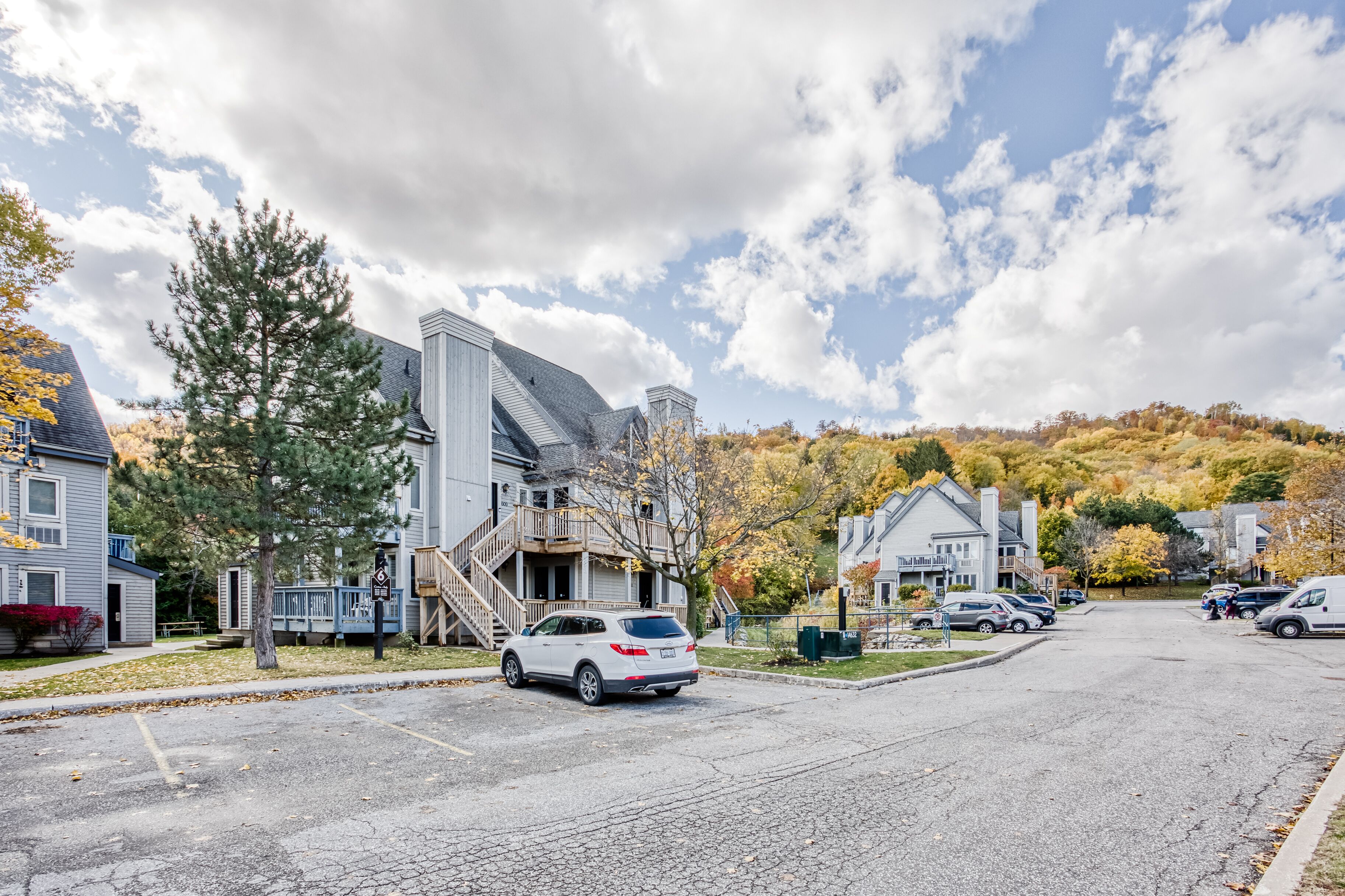 A residential street with houses, a white SUV parked on the side, and autumn foliage in the background.