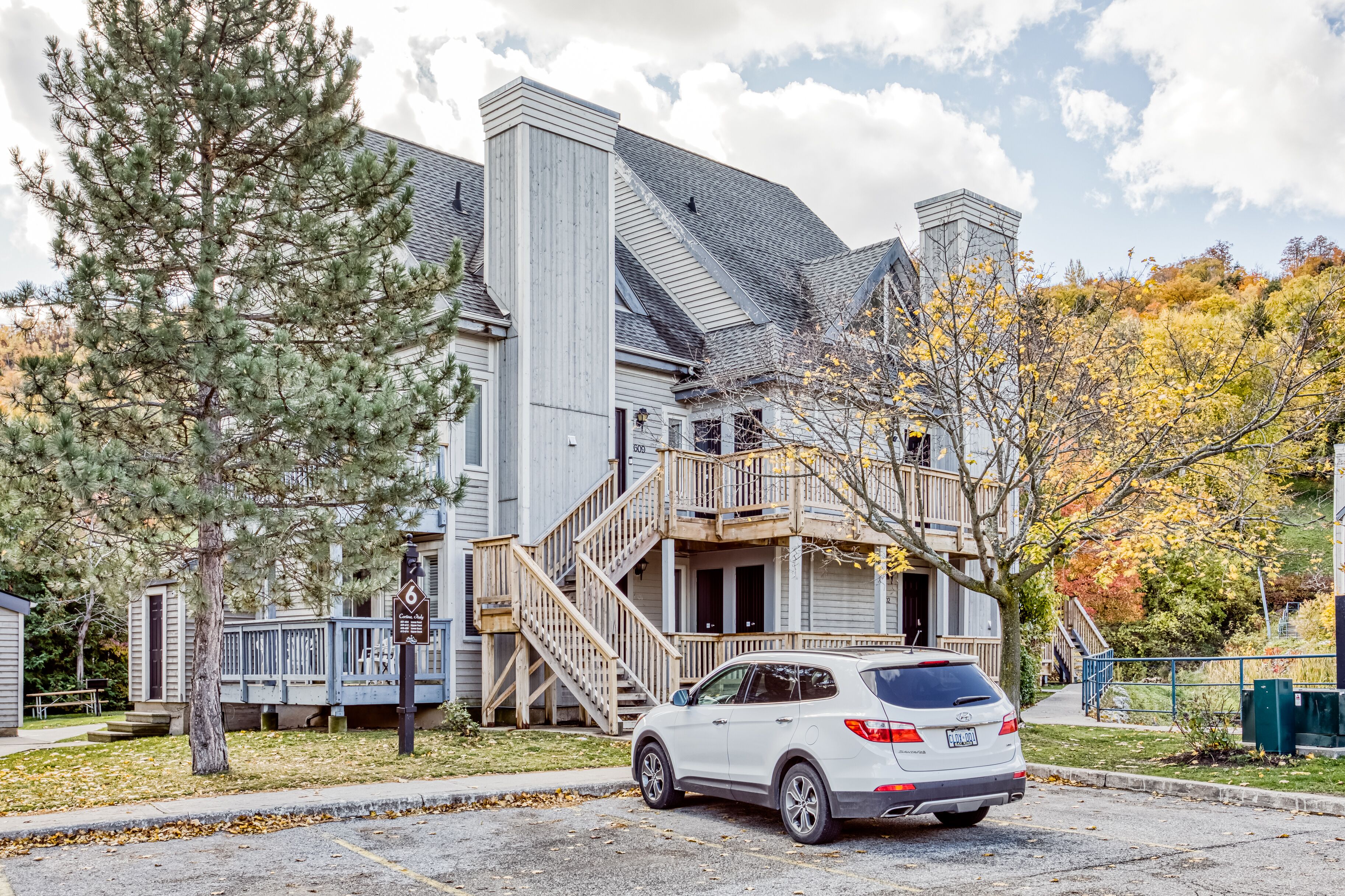 A two-story building with a staircase leading to a balcony, a parked white SUV, and a tree with autumn leaves.