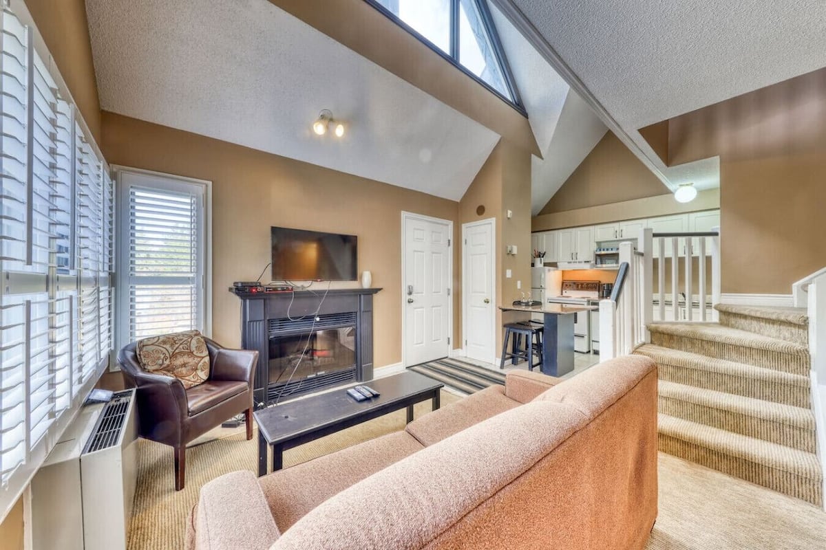 Una sala de estar con chimenea, un sofá, un televisor y una escalera que lleva a una cocina.