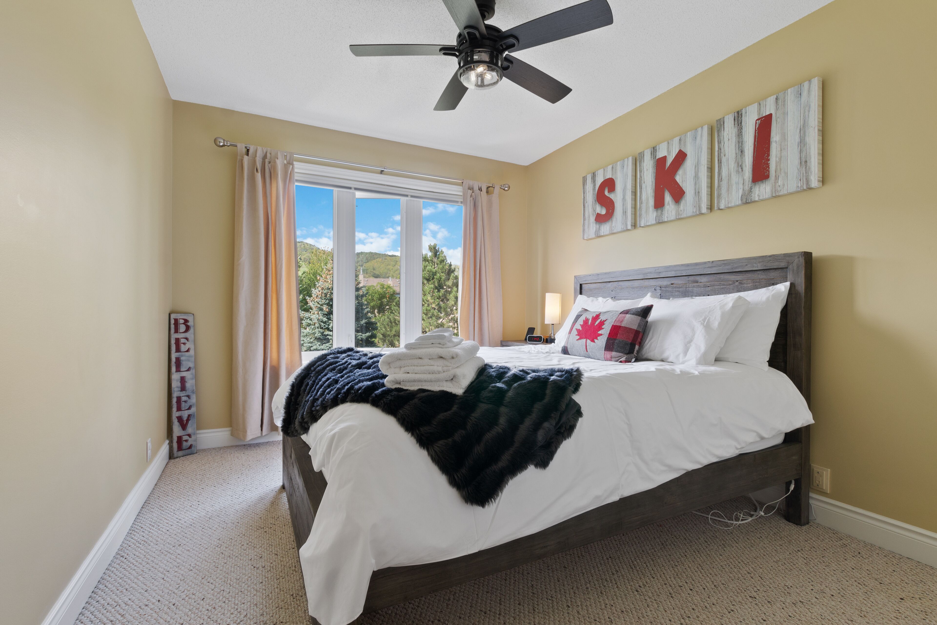 A bedroom with a bed, a ceiling fan, a window with curtains, and a wall with the word "SKI" spelled out.
