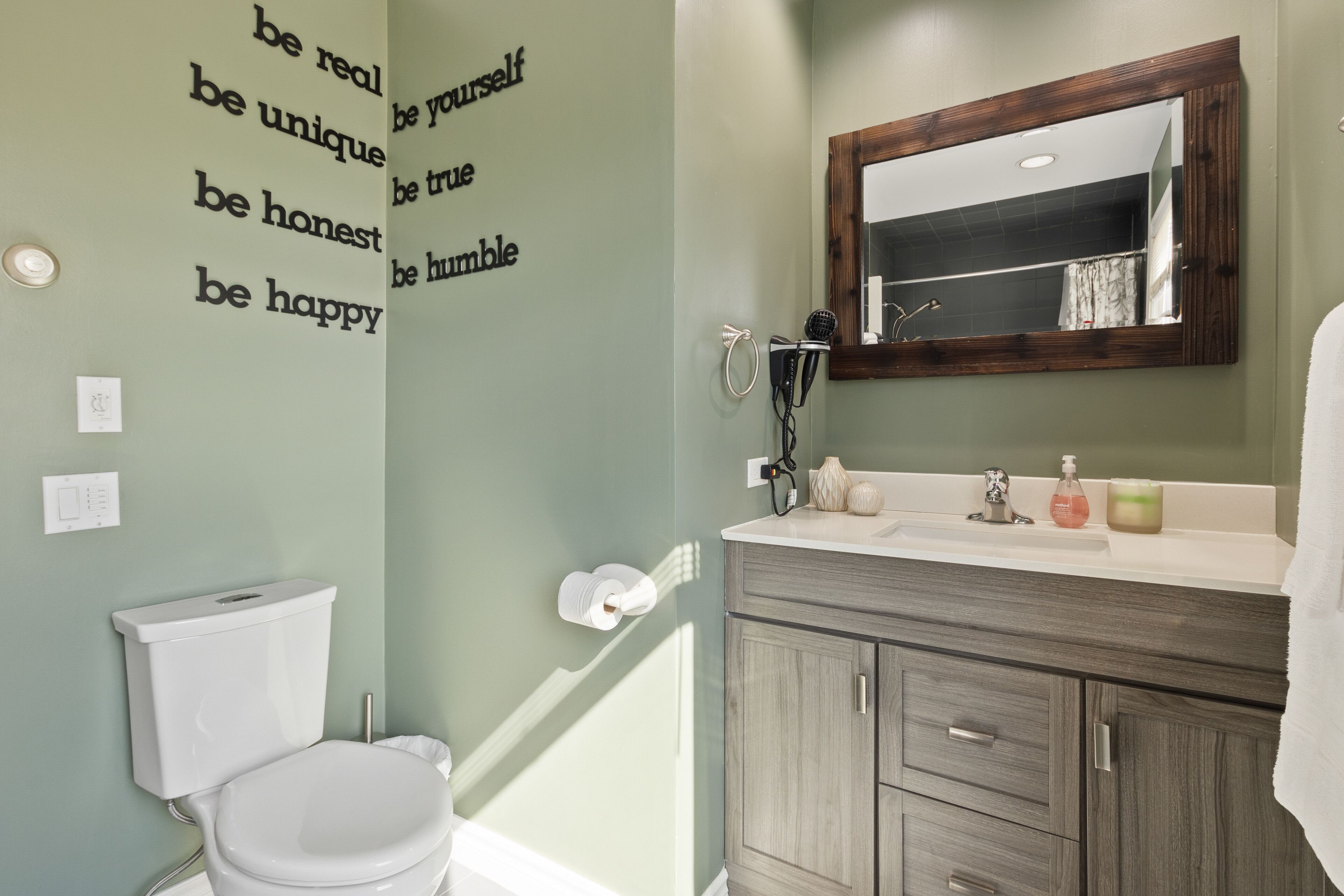 Un baño con inodoro, lavamanos y espejo. Hay citas motivacionales en la pared y artículos de aseo en el lavamanos.
