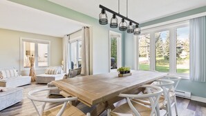 A spacious dining area with a wooden table and white chairs, large windows with curtains, and a view of greenery outside. - Sierra Lane Condos (The Blue Mountains)