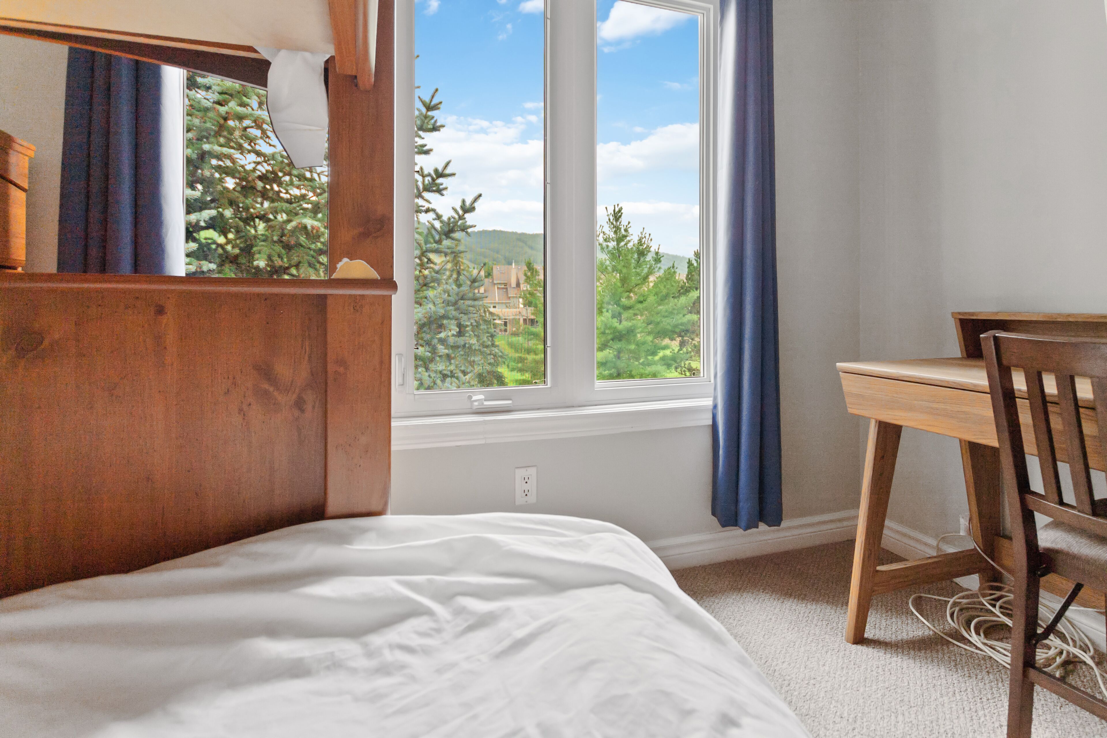 A bedroom with a bed, a wooden desk, and a window with a view of trees and hills.