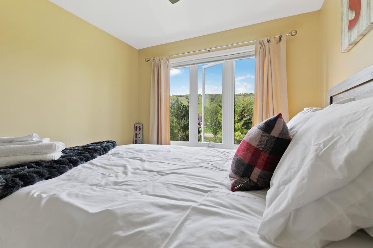 A bedroom with a bed, a window with curtains, a view of greenery outside, and a sign on the wall.