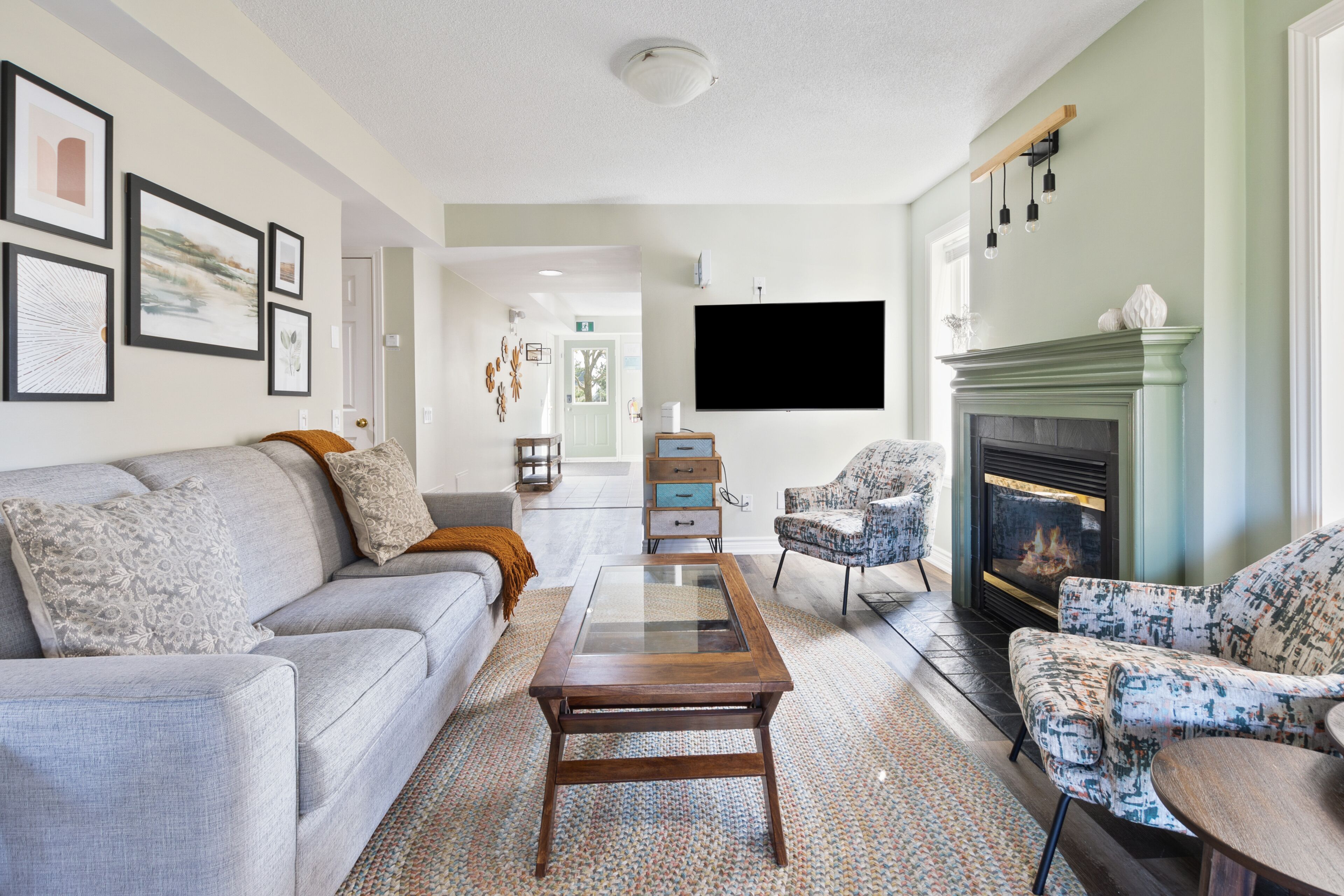 A modern living room with a sofa, armchairs, a glass-top coffee table, a fireplace, and a flat-screen TV.