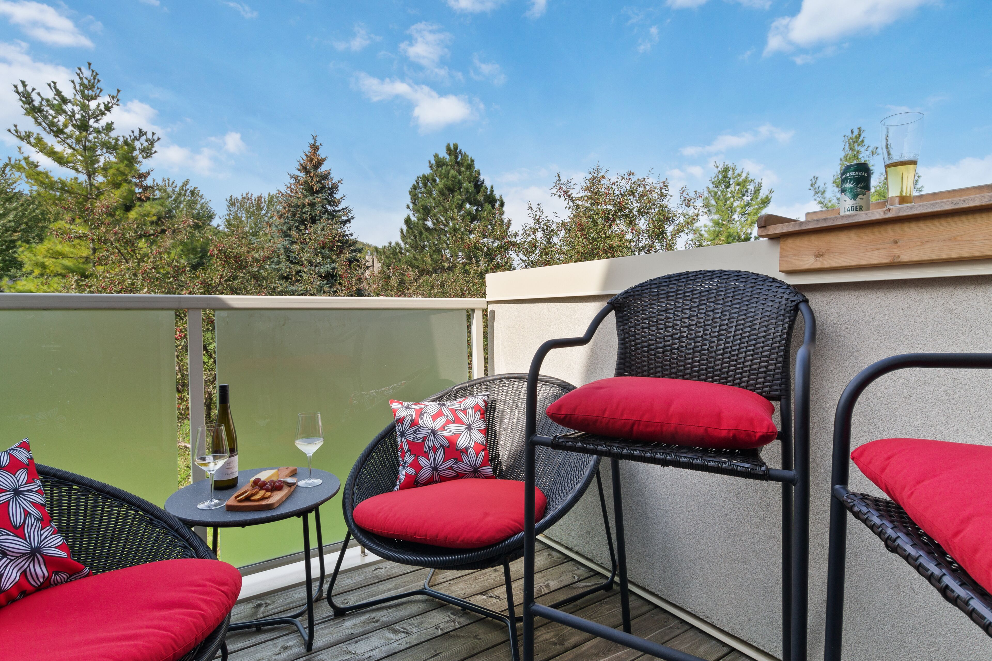 A balcony with two wicker chairs, each with red cushions, and a small table with a bottle of wine and glasses.