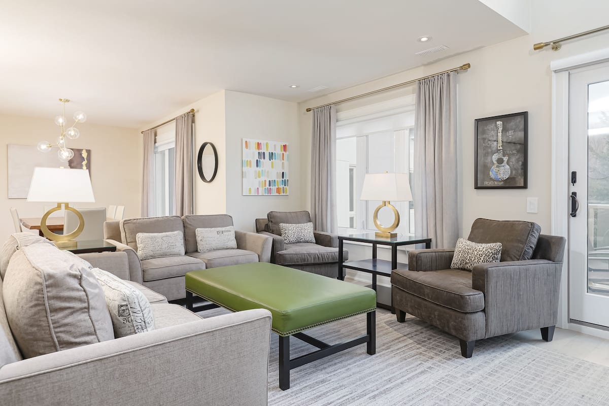 A modern living room with a green ottoman, grey sofas, and a coffee table. There are two lamps, a painting, and a framed item on the wall.