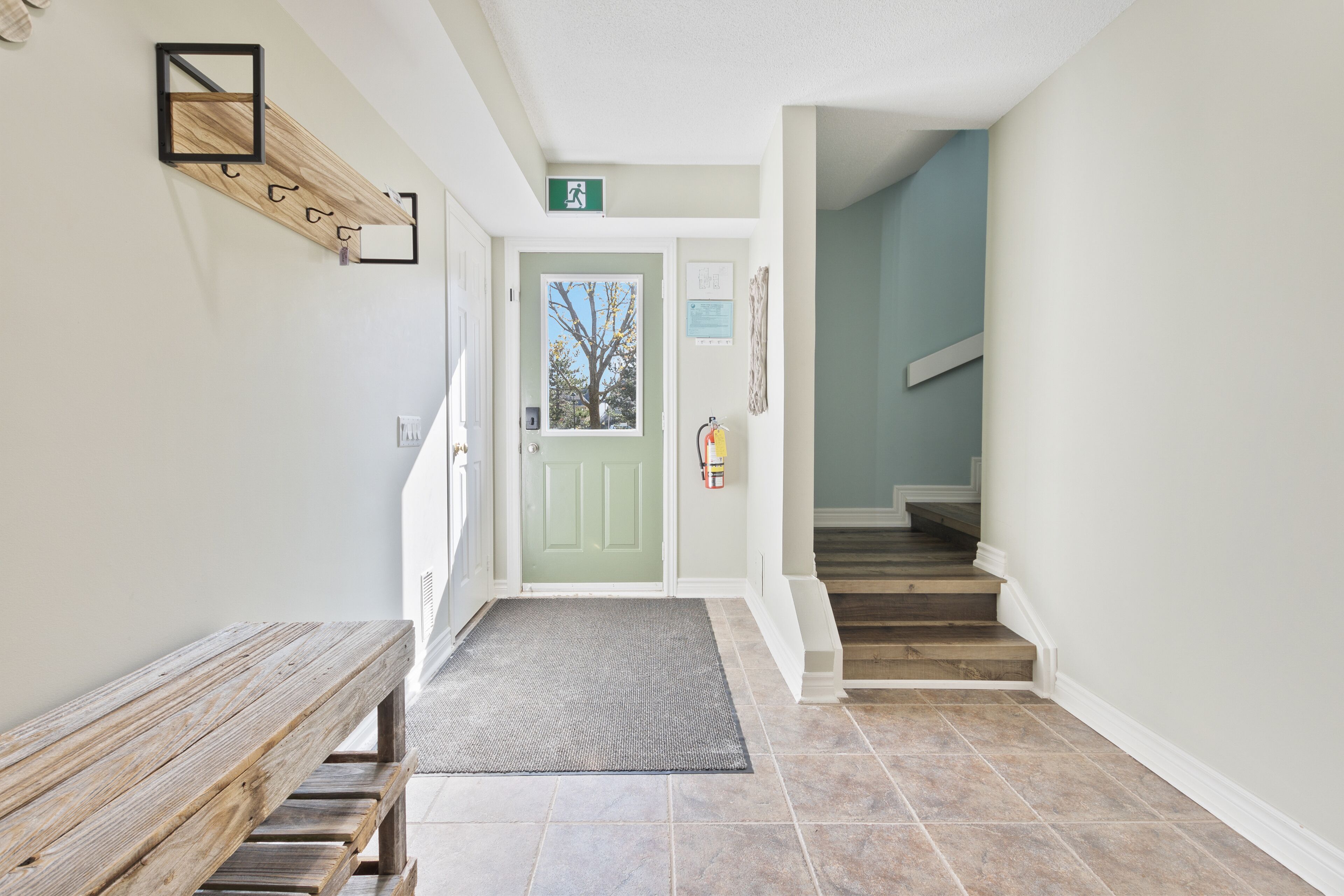Un pasillo limpio y moderno con un banco de madera, una puerta verde y una escalera que sube a otro piso.