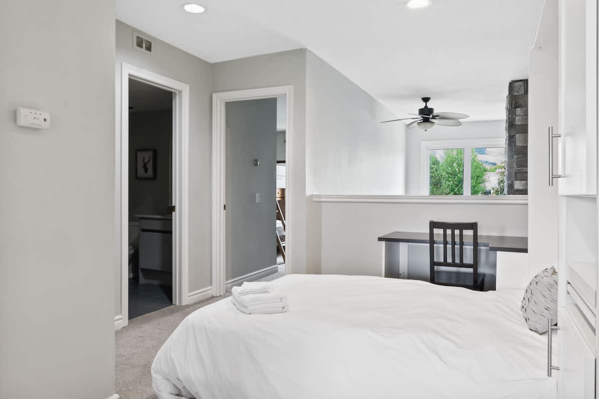 A bedroom with a bed, a desk, a chair, a ceiling fan, and a view of the outdoors.
