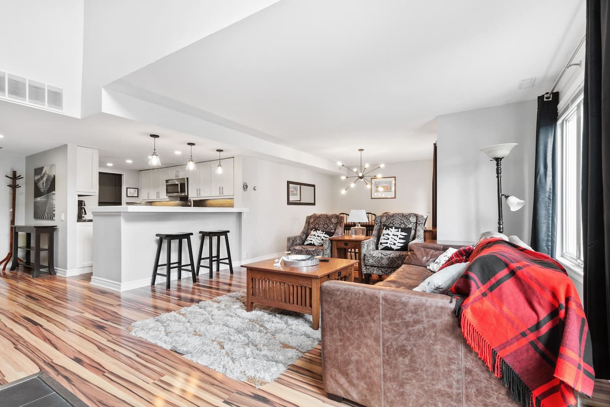 A modern living room with a sofa, coffee table, and a red plaid blanket. There is a kitchen area with bar stools and a dining table.