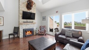 A living room with a stone fireplace, a flat-screen TV, a leather sofa, and a view through large windows. - Wintergreen Condos (The Blue Mountains)