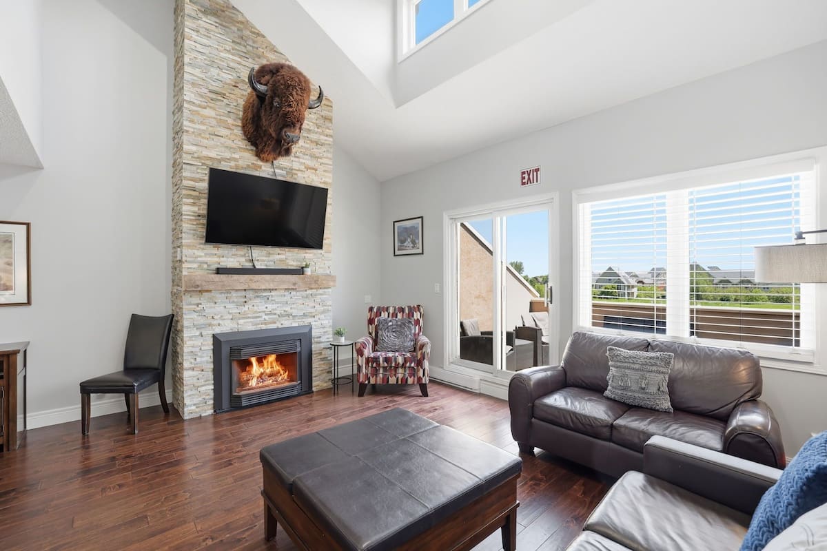 A living room with a stone fireplace, a flat-screen TV, a leather sofa, and a view through large windows.