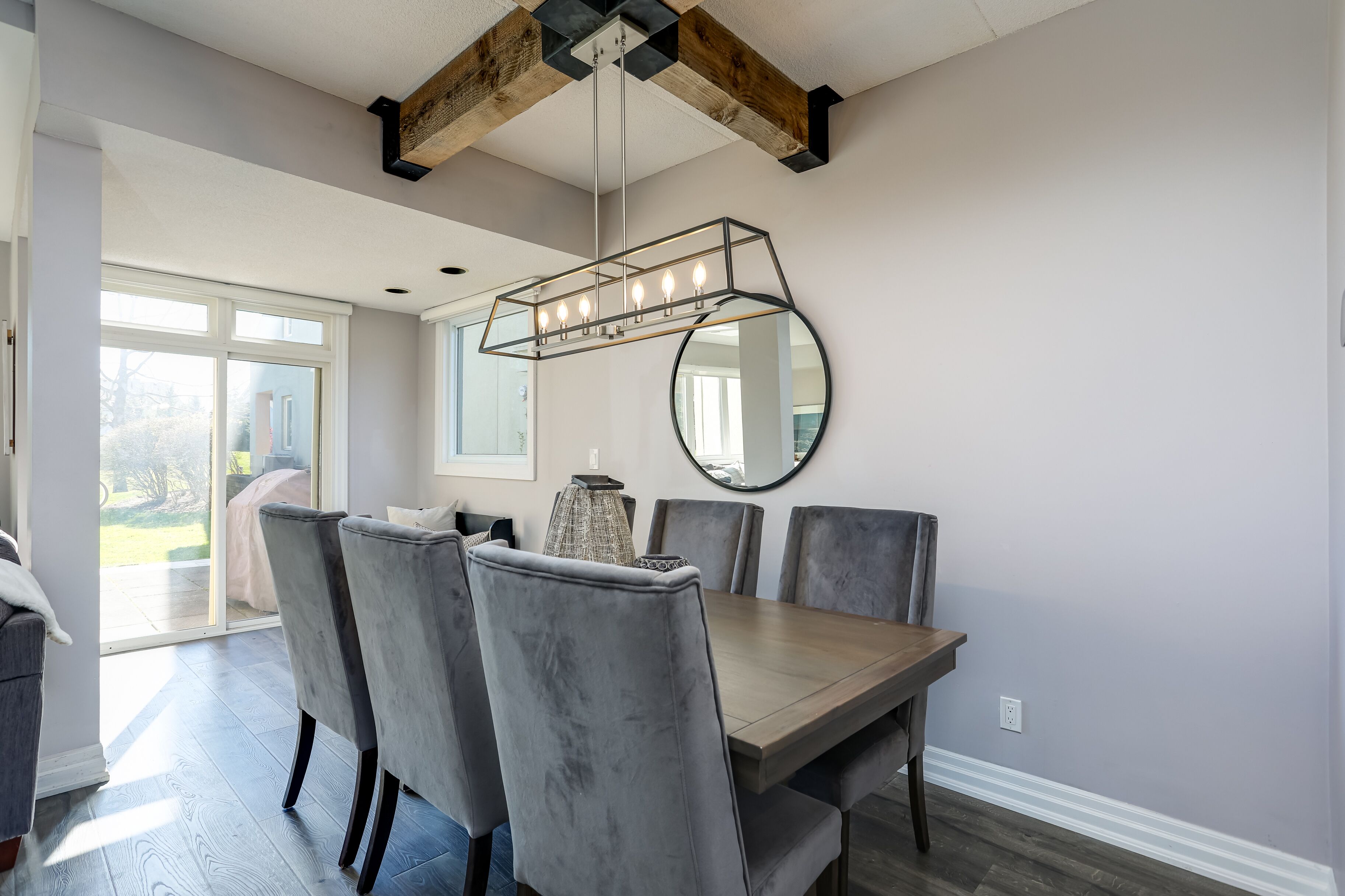 A dining area with a wooden table, grey upholstered chairs, a hanging light fixture, and a round mirror on the wall.