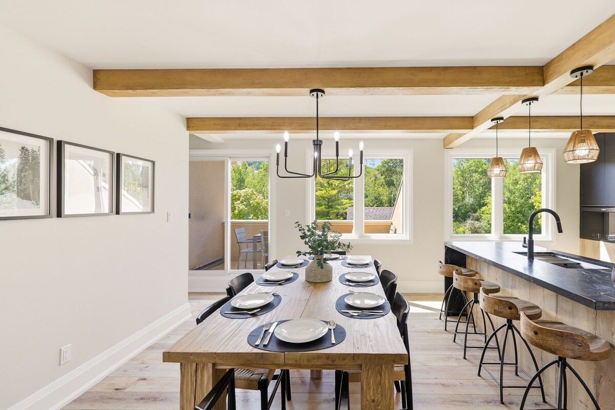 Un comedor con una larga mesa de madera dispuesta para seis, una cocina con encimera negra y vistas al exterior con vegetación.