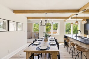 Un coin repas avec une longue table en bois dressée pour six personnes, une cuisine dotée d’un plan de travail noir et une vue sur la verdure à l’extérieur.