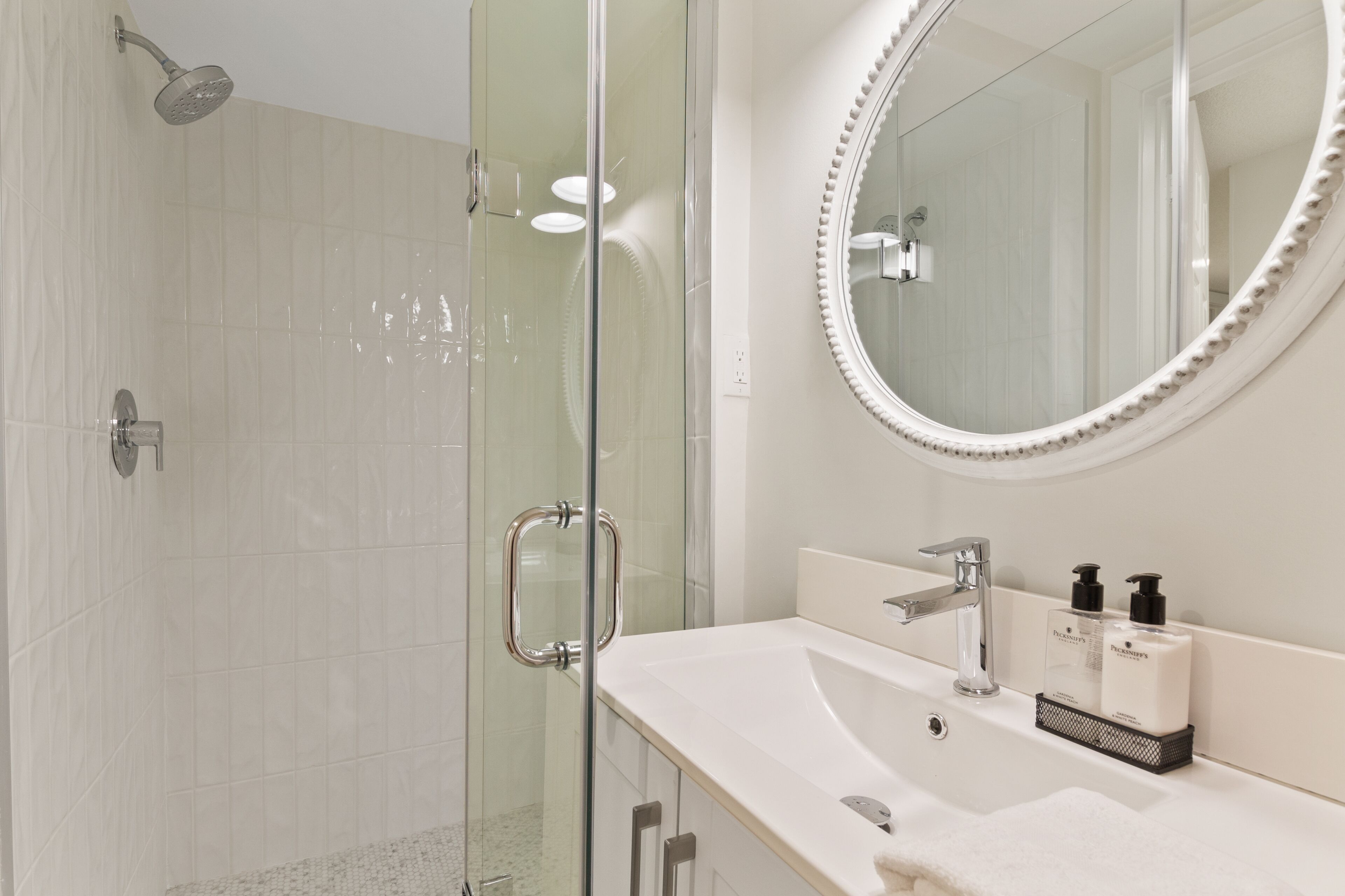 A modern bathroom with a glass shower enclosure, a white sink with a round mirror, and two bottles of shampoo on the counter.