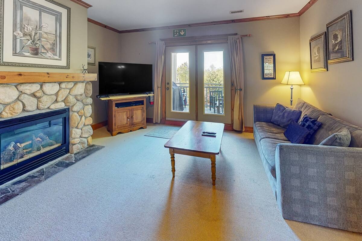 Ein Wohnzimmer mit Kamin aus Stein, einem Flachbildfernseher, einem Holztisch, einem grauen Sofa und einer Balkontür.