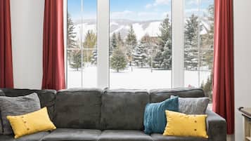 A grey sofa with yellow and blue cushions, a wooden coffee table, and a view of snow-covered trees and mountains through large windows.