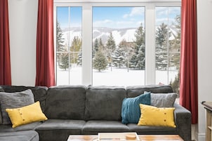 Un sofá gris con cojines amarillos y azules, una mesa de centro de madera y vistas a árboles y montañas nevadas a través de grandes ventanales.