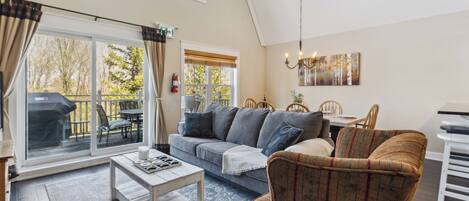 A spacious living room with a large sliding glass door leading to a deck, a sofa set, a coffee table, and a dining area with a table and chairs.
