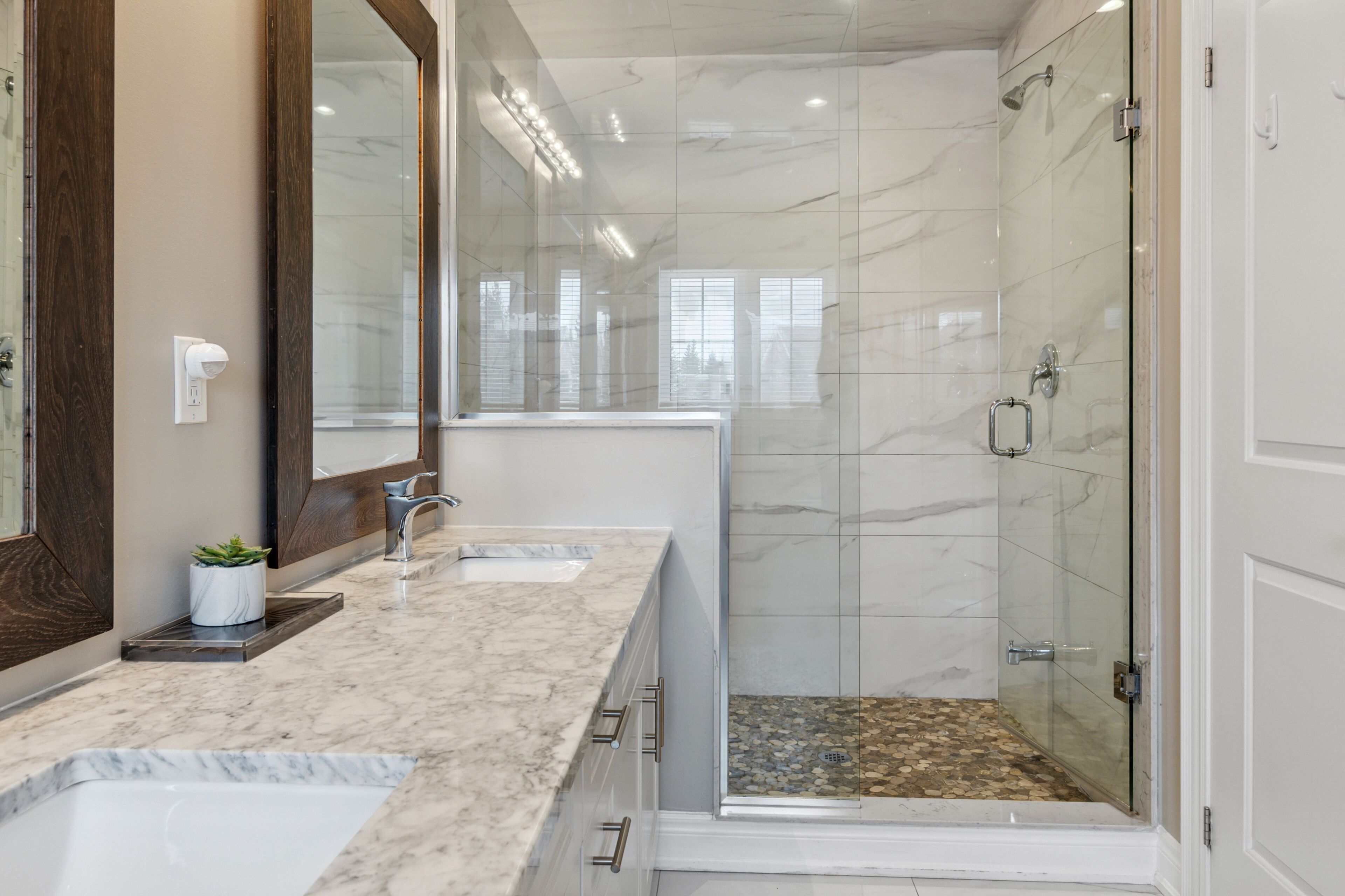 A modern bathroom with a marble countertop, a large mirror, a glass shower enclosure, and a potted plant.