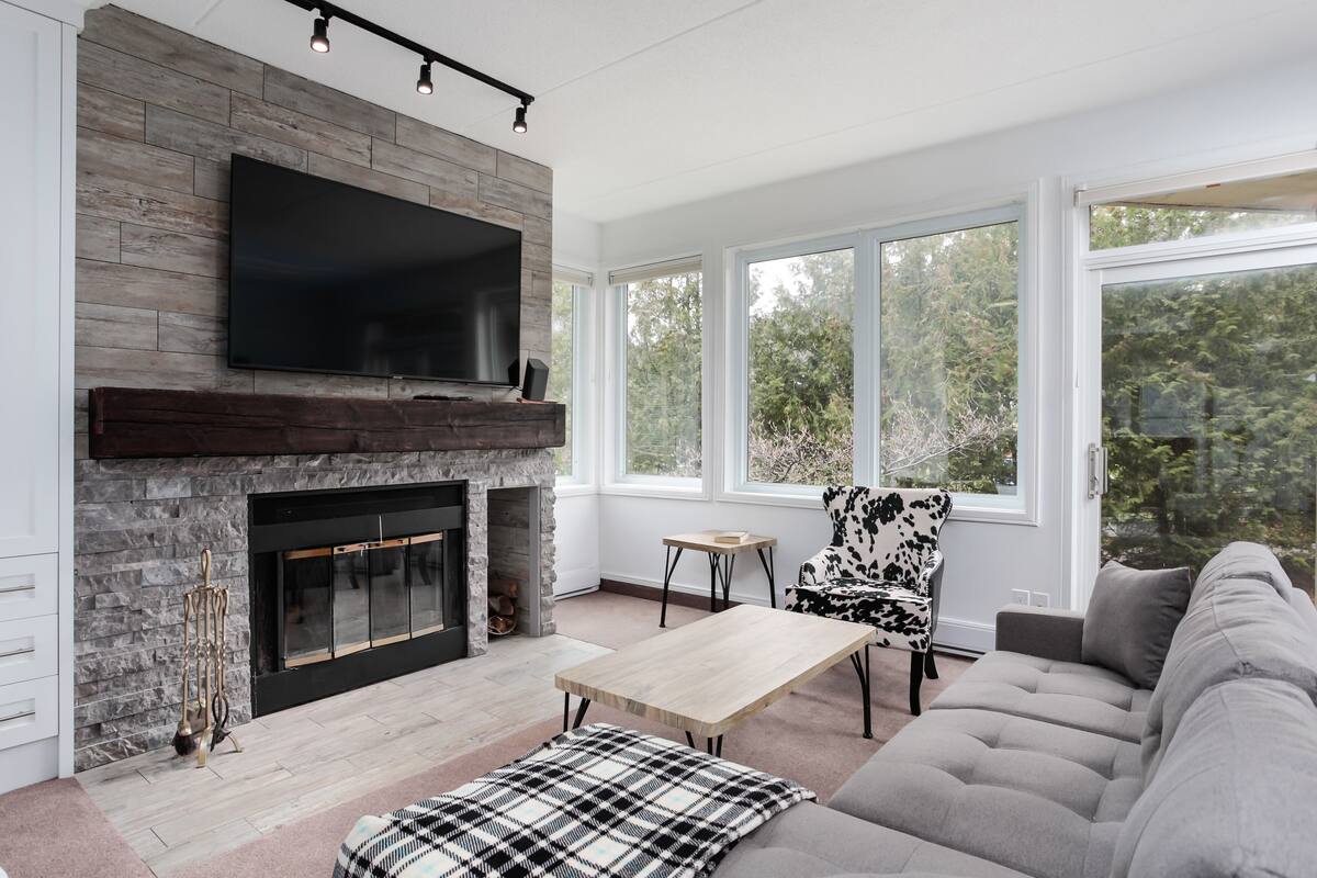 A modern living room with a fireplace, a flat-screen TV mounted above it, a large window offering a view of trees, a sofa, and a coffee table.